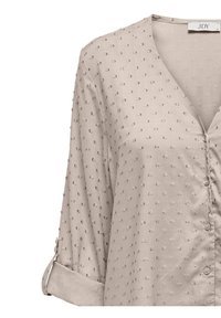 Blusa beige chiaro con scollo a V, chiusura frontale a bottoni e maniche arrotolabili. Presenta un tessuto testurizzato con piccoli punti decorativi distribuiti uniformemente.