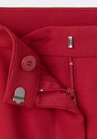 Gros plan de la ceinture d’un vêtement rouge montrant un bouton, une fermeture éclair, un crochet en tissu et une boutonnière sur un tissu texturé.