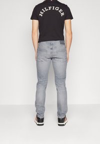 Camiseta negra con el logo "HILFIGER" en la espalda, combinada con jeans gris claro y zapatillas negras con detalles en rojo. Diseño minimalista, look casual.