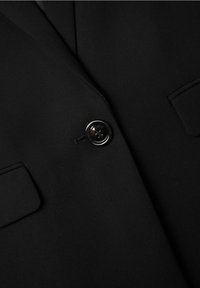 Tissu de blazer noir avec une texture lisse, comportant un seul bouton noir et deux poches poitrine sans motifs ni ornements visibles.