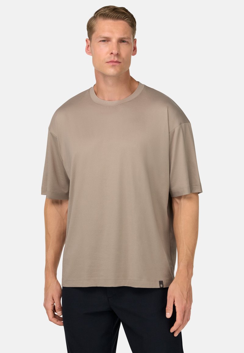 Béžové oversized tričko s kulatým výstřihem a krátkými rukávy, vyrobené z měkkého materiálu, s malou nášivkou loga na spodním lemu.