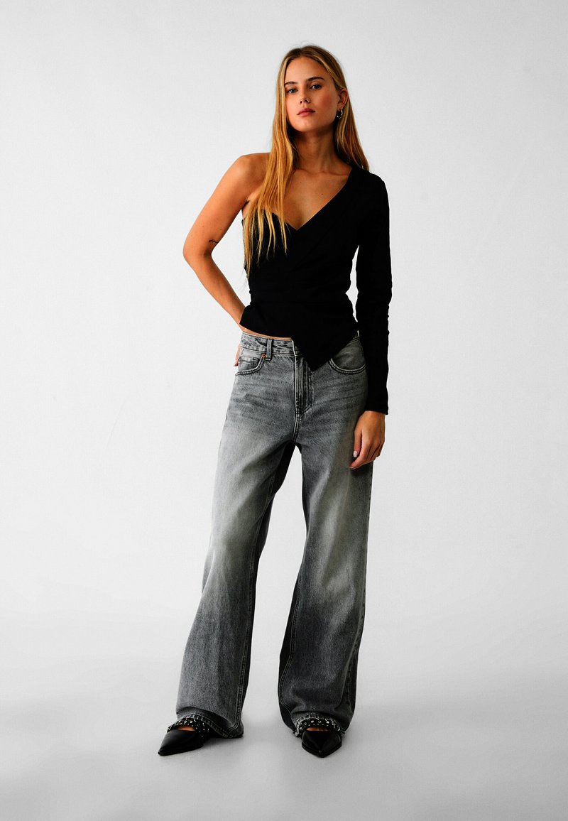 Top nero a spalla unica con un design drappeggiato, abbinato a jeans grigi chiari a gamba larga. La modella indossa delle ballerine nere a punta, rivelando un look elegante.