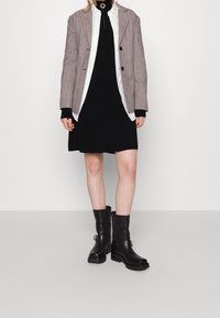 Frau in schwarzem Kleid, weißem Hemd, grauem Blazer und schwarzen Lederstiefeln bis zur Mitte der Wade, die vor einem schlichten Hintergrund steht.