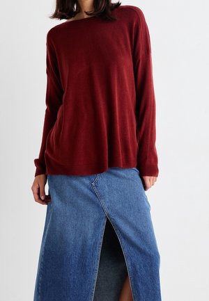 Vrouw draagt een losse, lange mouwen bordeauxrode trui gecombineerd met een blauwe denim rok met een centrale voorste split.