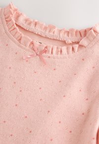 Hele roosa puuvillane kampsun volangidega kaelusega, täpilise tekstuuriga ja väikese roosa paelaga kaelusel.