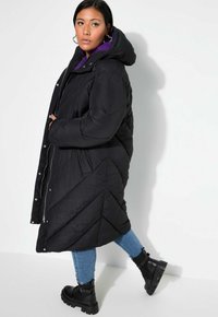 Svart puffjacka med huva, med ett chevron-quiltat mönster, framtida dragkedja och tryckknappar. Bärs över en lila topp och denimjeans.