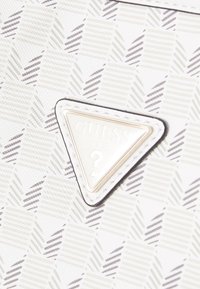 Gros plan sur un tissu à motifs blancs et beiges avec un patch triangulaire blanc du logo Guess U.S.A. cousu dessus.
