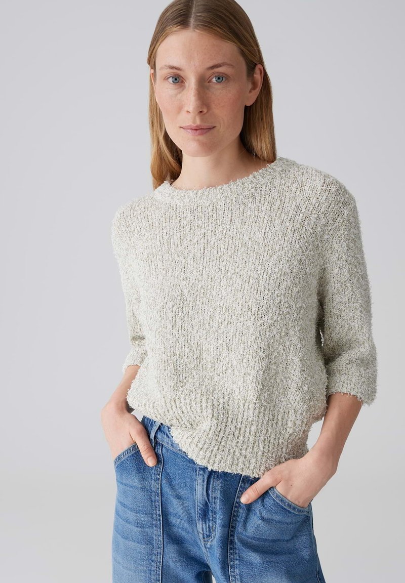 Frau mit glatten blonden Haaren, die einen strukturierten hellgrauen Pullover und eine blaue Jeans trägt, mit den Händen in den Taschen, neutraler Hintergrund.