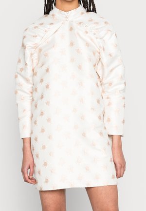 Frau trägt ein weißes Satin-Minikleid mit aufgepolsterten Schultern und hellrosa Blumenmustern, stehend vor einem schlichten Hintergrund.