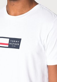 Biała bawełniana koszulka z okrągłym dekoltem, z granatowym prostokątnym logo z białymi i czerwonymi akcentami. Krótkie rękawy i gładka faktura.