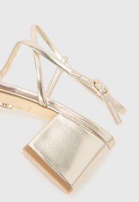 Goldene metallische Plattform-Sandale mit Blockabsatz, ausgestattet mit überkreuzten Riemen und verstellbarem Knöverschluss. Glatte Textur und glänzende Oberfläche.