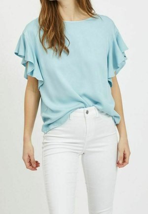 Blouse bleu clair à manches courtes avec manches à volants superposés portée avec un pantalon blanc taille haute, visible du cou aux hanches.