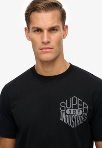 Camiseta de algodón negra con cuello redondo; presenta un gráfico hexagonal en gris que dice "SUPER DRY INDUSTRIES" en el lado izquierdo del pecho.