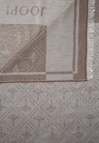 Textile beige à motifs avec des formes de diamant et de petits motifs floraux, montrant un coin replié avec une frange sur un bord.