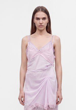 Jeune femme aux cheveux bruns lisses porte une robe légère rose pâle transparente avec des détails en dentelle et de fines bretelles doubles.