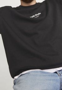 Uomo che indossa una maglietta nera con testo bianco "Studio" e jeans azzurri chiari, tagliati al petto e in vita, su uno sfondo chiaro e semplice.