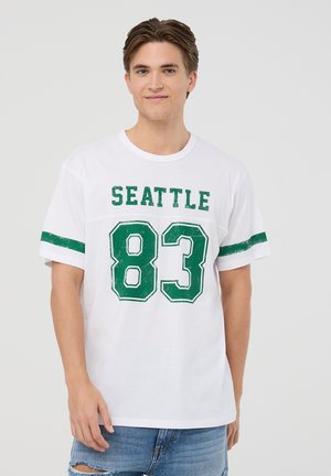 T-shirt bianco in cotone con testo verde "SEATTLE" e stampa grafica "83". Strisce verdi orizzontali sulle maniche, vestibilità rilassata, design a girocollo.