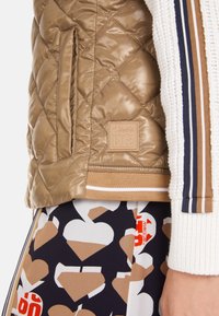 Quilted, glossy tan jacket with side pockets, ribbed waistband, and logo patch. Paired with patterned skirt featuring hearts and contrasting colors.