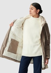 Giacca puffer color beige con zip frontale, foderata internamente in crema e con una tasca. Indossata sopra un maglione lavorato a maglia color crema e pantaloni blu scuro.
