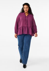 Top peplum prugna con maniche lunghe, scollo a V e dettaglio con cordino, abbinato a jeans blu a gamba dritta e stivaletti neri.