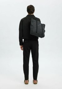 Schwarze Tasche aus robustem Stoff mit doppelten schwarzen Trägern. Die Person trägt ein schwarzes Shirt und schwarze Hosen und steht vor einem hellgrauen Hintergrund.