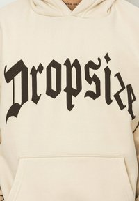 Beige hoodie with a front pocket and a large black gothic-style print reading "Dropsize." Soft texture, casual design, and hooded style.