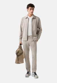Lys grå zip-up jakke over en hvid polo, matchende grå bukser og en beige rygsæk. Sneakers har et beige og marineblåt design.