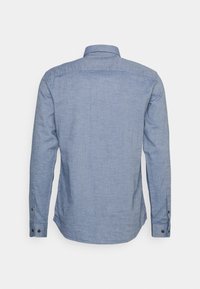 Camicia in chambray azzurro chiaro a maniche lunghe con polsini abbottonati, vista da dietro su uno sfondo bianco semplice.