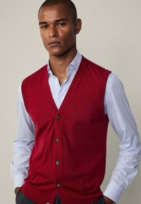 Gilet rosso lavorato a maglia con scollatura a V, dotato di cinque bottoni scuri. Indossato sopra una camicia a righe blu chiaro con colletto abbottonato.