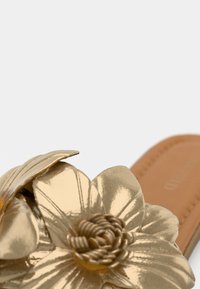 Κοντινή λήψη μεταλλικού λουλουδιού σε χρυσό χρώμα, λεπτομέρεια σε λουράκι δερμάτινου σανδάλι σε ταμπά απόχρωση, σε λευκό φόντο.