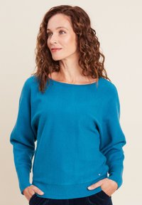 Turkiser Strickpullover mit weitem Ausschnitt, lockerer Passform und gerippten Ärmeln. Auffälliges Detail: kleines Logo-Accessoire an der Hüfte.