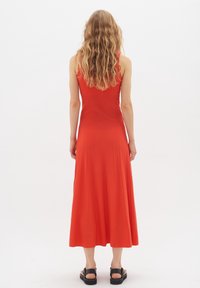 Červené maxi šaty bez rukávů s přiléhavým živůtkem a rozšířenou sukní, s žebrovanou strukturou a jemnými skladbami, doplněné černými plochými sandály.