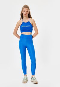 Blauwe sportbeha met het woord "BRIGHT" in het wit, gecombineerd met bijpassende blauwe high-waisted leggings. Model draagt neutraalgekleurde sneakers.