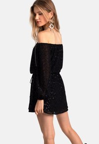 Robe noire décolletée avec un tissu transparent texturé, des manches élastiques et une taille à cordon, ornée de motifs floraux subtils.