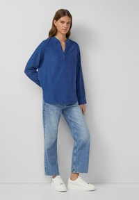 Chemise bleue à manches longues avec col à notch, assortie à un jean en denim clair et des baskets blanches. Le tissu a une texture douce.