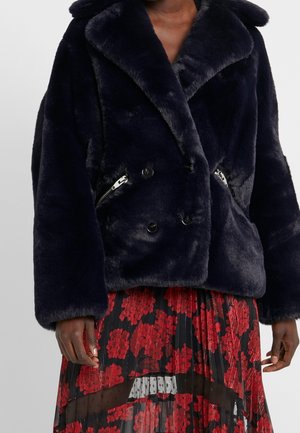 Tummansininen keinoturkistakki, jossa on kaksirivinen napitus ja hopeanväriset vetoketjutaskut, pidetty mustan ja punaisen kukkakuosisen läpikuultavan pliseeratun hameen päällä.