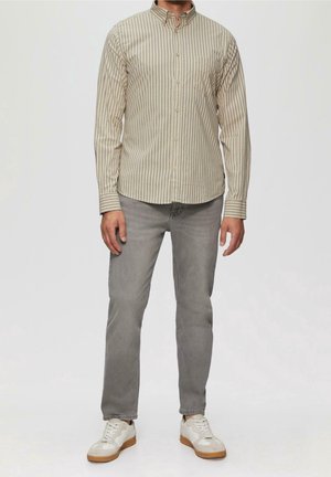 Man draagt beige gestreept overhemd met knopen, grijze spijkerbroek en witte sneakers, staand tegen een effen achtergrond.