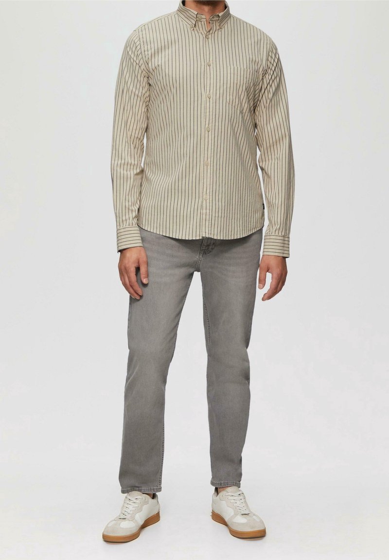 Homme portant une chemise beige rayée à boutons, un jean gris et des baskets blanches, debout devant un fond uni.