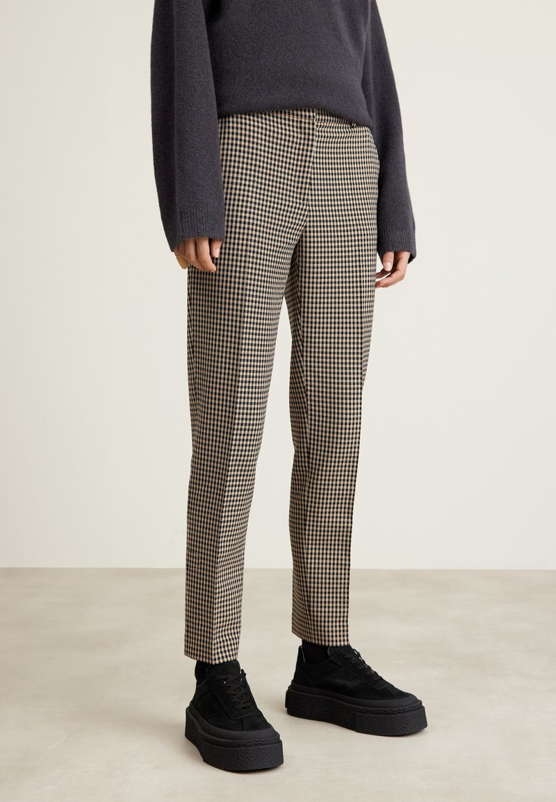 Personne portant un pull noir, un pantalon à motif pied-de-poule beige et noir, et des baskets compensées noires, debout sur un sol clair.