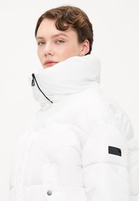 Witte pufferjack met hoge kraag, ritssluiting en zwarte accenten. Kenmerken zijn een gestructureerde afwerking en zijzakken met drukknoopsluitingen.