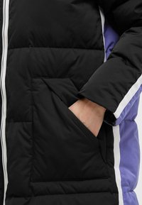 Schwarze Steppjacke mit einer großen Fronttasche, weißen Akzentstreifen und einem kontrastierenden lila Seitenpanel. Glatte Textur, gepolstertes Design.