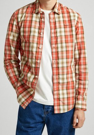 Homme portant une chemise à carreaux rouge, beige et blanche déboutonnée, par-dessus un t-shirt blanc et un jean bleu, avec une main dans la poche.