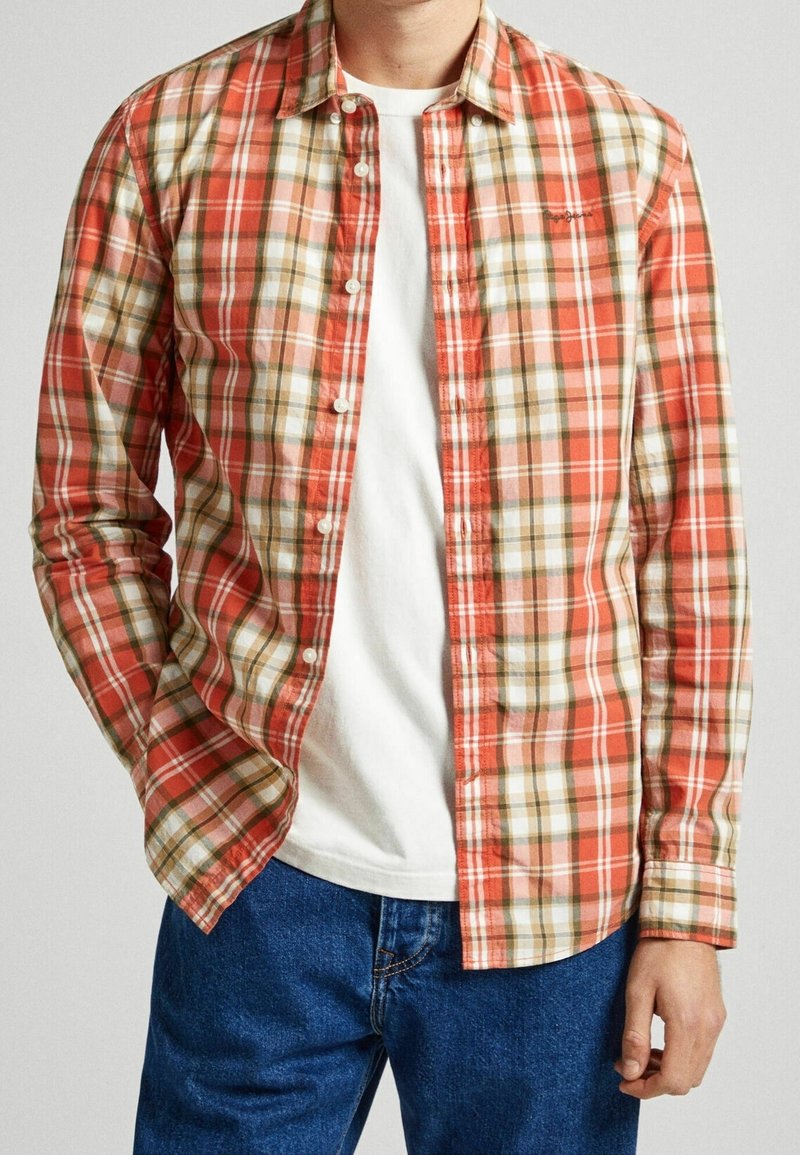 Homme portant une chemise à carreaux rouge, beige et blanche déboutonnée, par-dessus un t-shirt blanc et un jean bleu, avec une main dans la poche.