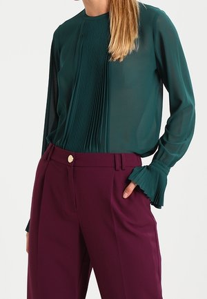 Frau trägt eine dunkelgrüne, plissierte Bluse mit langen Ärmeln und ausgestellten Manschetten sowie hoch taillierte violette Hose mit Knopfverschluss.