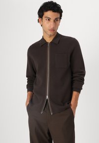 Cardigan maro cu fermoar, cu guler, textură tricotată și un buzunar pe piept. Prezintă un fermoar argintiu și mâneci lungi. Purtați-o cu pantaloni maro asortați.