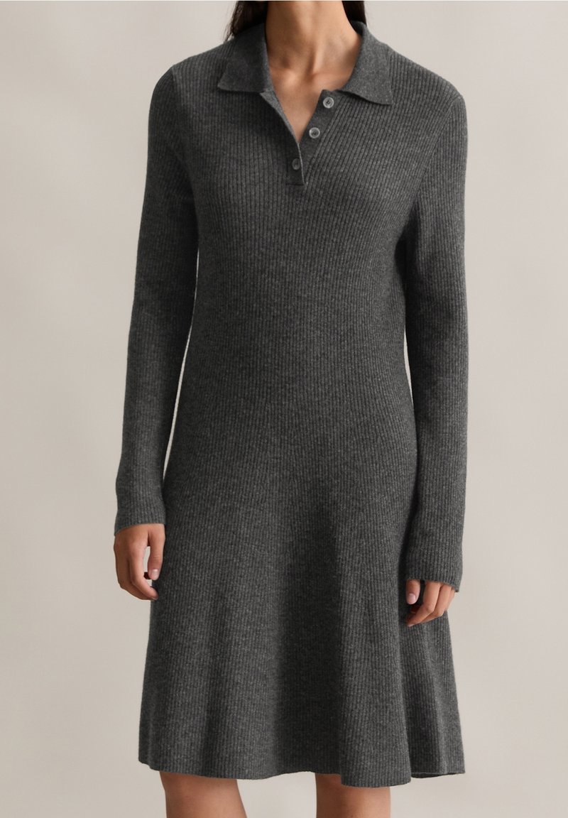 Femme portant une robe en maille côtelée à manches longues, de couleur gris foncé, avec un col et trois boutons au niveau de l'encolure, debout devant un fond uni.