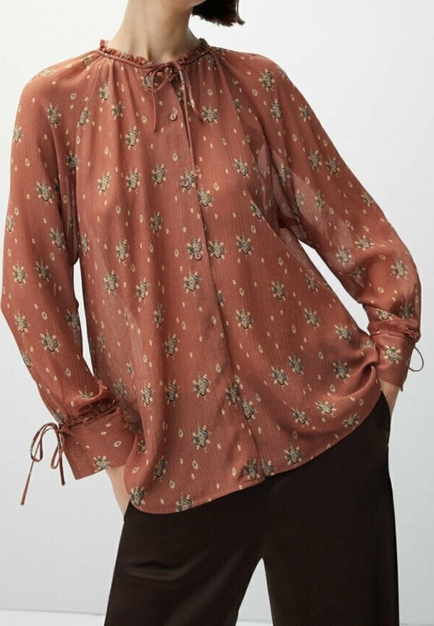 Blusa a maniche lunghe color ruggine realizzata in tessuto leggero, con motivi floreali, scollatura arricciata con lacci e polsini elasticizzati.