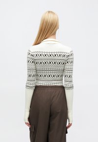 Adīts džemperis ar baltu pamatu un brūniem ģeometriskiem rakstiem, garām piedurknēm un apkakli; kombinēts ar brūnām, platām biksēm.