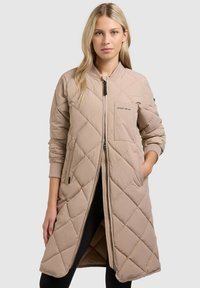 Femeie blondă purtând o geacă lungă bej matlasată cu fermoar frontal, stând cu o mână în buzunar pe un fundal simplu.