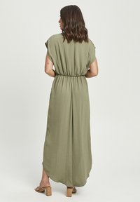 Woman with shoulder-length hair wearing an olive green, short-sleeve, ankle-length dress and tan block-heeled sandals, facing away.